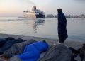 Migrants and refugees sleep as a man from Pakistan covered with a blanket looks on a ferryboat leaving the port of the Greek island of Chios, Wednesday, April 6, 2016.Volunteers are concerned about children health of some 300 migrants and refugees that manage to leave the VIAL detention center a few days ago. Authorities said that more than 1700 migrants and refugees are in the island. (AP Photo/Petros Giannakouris)
