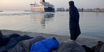 Migrants and refugees sleep as a man from Pakistan covered with a blanket looks on a ferryboat leaving the port of the Greek island of Chios, Wednesday, April 6, 2016.Volunteers are concerned about children health of some 300 migrants and refugees that manage to leave the VIAL detention center a few days ago. Authorities said that more than 1700 migrants and refugees are in the island. (AP Photo/Petros Giannakouris)