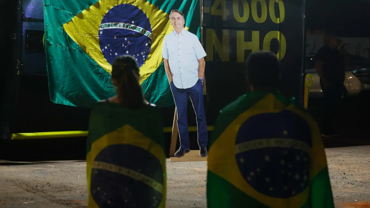 Una foto notturna con persone che indossano bandiere brasiliane come mantelli, le loro spalle sono rivolte alla telecamera mentre sono in piedi davanti a un grande ritaglio di cartone a grandezza naturale del politico brasiliano Jair Bolsonaro. Dietro il ritaglio, è esposta una grande bandiera del Brasile