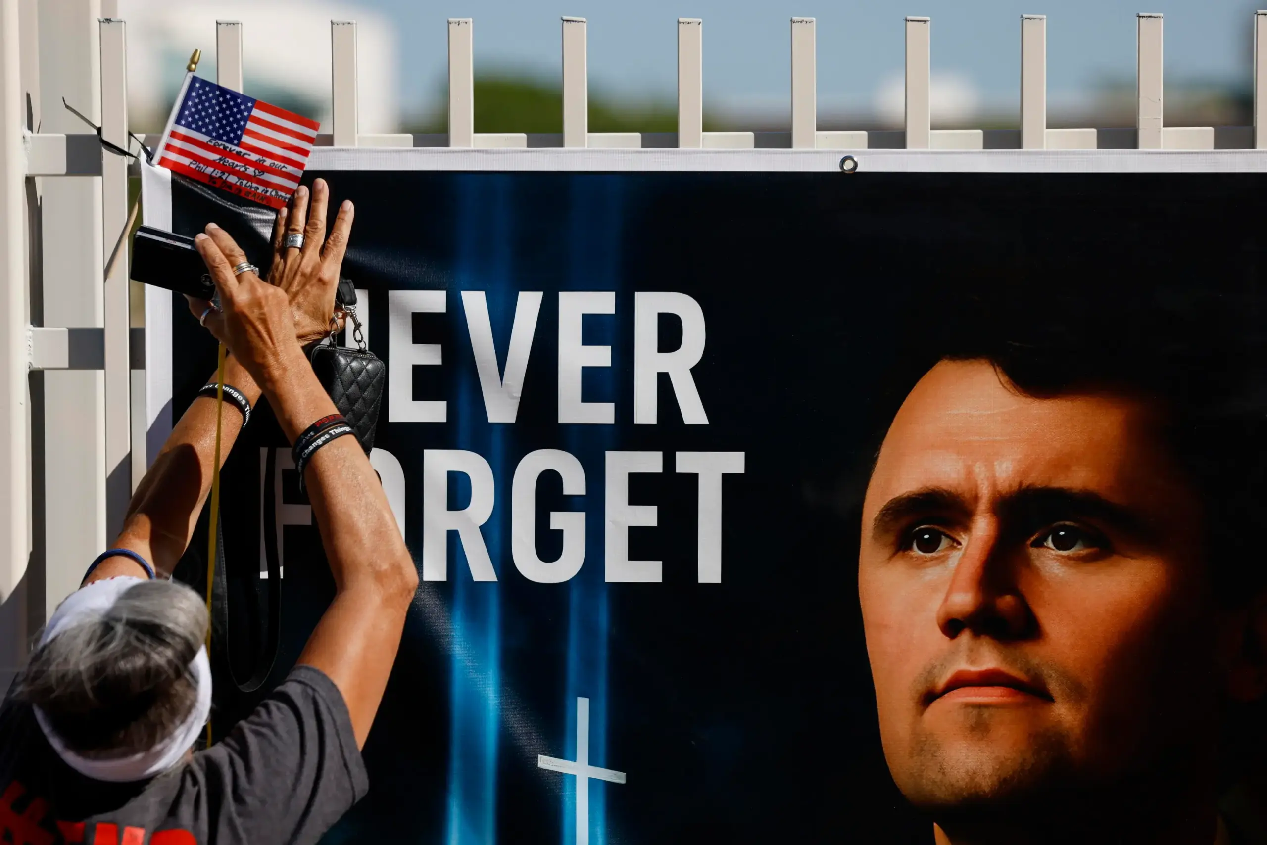 Una persona con una maglietta grigia e una visiera bianca alza le mani per attaccare una piccola bandiera americana a un cartellone commemorativo improvvisato che recita 'NEVER FORGET' con una grande immagine del volto dell'attivista politico conservatore Charlie Kirk. Il memoriale è allestito su una recinzione all'esterno della sede centrale di Turning Point USA a Phoenix, Arizona