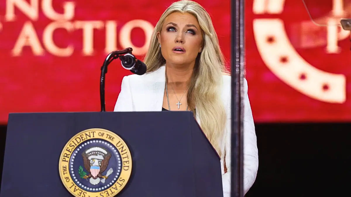 Erika Kirk,vedova di Charlie Kirk, con i capelli biondi e un blazer bianco, tiene un discorso con un'espressione commossa al microfono. Si trova dietro un podio blu scuro che reca il sigillo del Presidente degli Stati Uniti, in una sala con uno sfondo rosso sfocato dello State Farm Stadium di Glendale, Arizona, USA, dove si tiene la cerimonia funebre pubblica dell'attivista politico