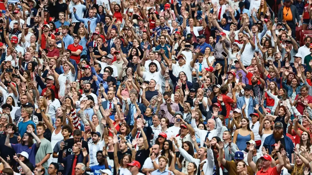 Una folla numerosa di persone, molte delle quali indossano vestiti rossi, bianchi e blu, che alzano le mani in aria in segno di acclamazione durante un evento la cerimonia commemorativa pubblica dell’attivista politico Charlie Kirk allo State Farm Stadium di Glendale, Arizona, USA