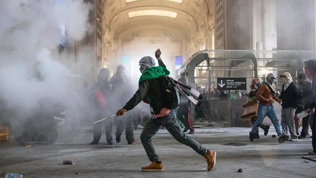 L'immagine cattura un momento di intensa tensione durante una protesta pro-Palestina a Milano. Lo scenario è l'ingresso della Stazione Centrale, visibile sullo sfondo.
Un manifestante, con il volto coperto da una kefiah, è in primo piano mentre lancia un oggetto. Intorno a lui c'è del fumo, probabilmente causato da un lacrimogeno o un fumogeno, e si vedono altri manifestanti che si muovono nel caos. I detriti a terra, come sassi e pezzi di barricate, testimoniano la violenza degli scontri tra i manifestanti e la polizia