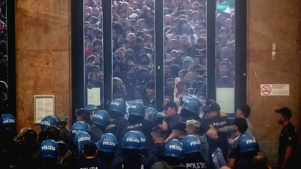 L'immagine mostra un gruppo di agenti di polizia in tenuta antisommossa, visti di spalle, che fronteggiano una folla molto fitta che preme contro una grande vetrata.
La scena rappresenta un momento di tensione, con gli agenti che formano una barriera. Oltre la vetrata, i volti e le sagome della gente sono ammassati, creando un'impressione di pressione e di affollamento estremo. L'immagine cattura il contrasto tra la linea ordinata della polizia e il caos della folla che cerca di superare la barriera di vetro.