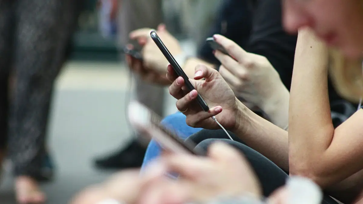 Una vista ravvicinata mostra diverse persone giovani sedute, la maggior parte delle quali sta tenendo e guardando i propri smartphone. Le loro mani e i dispositivi sono in primo piano, con figure sfocate sullo sfondo