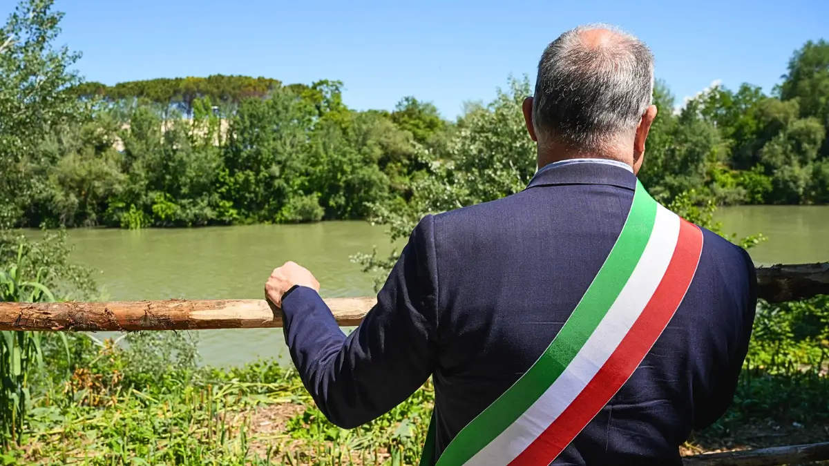 Il sindaco di Roma Roberto Gualtieri, visto di spalle, indossa un abito blu scuro con la fascia tricolore verde, bianca e rossa. È appoggiato a una staccionata di legno e guarda verso il fiume Tevere, che scorre placido in mezzo alla vegetazione rigogliosa. Sullo sfondo, alberi e pini marittimi si stagliano sotto un cielo limpido. L’occasione è l’inaugurazione del Parco fluviale all’Acqua Acetosa, il 10 maggio 2025