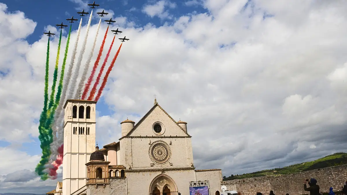 Una squadriglia di aerei a reazione, le Frecce Tricolori, sta sorvolando il cielo azzurro e nuvoloso. Lasciano dietro di sé strisce di fumo nei colori della bandiera italiana: verde, bianco e rosso. Gli aerei sono in formazione, con alcuni che volano più in alto e altri più in basso. Sotto di loro si trova la facciata e il campanile di una grande chiesa di pietra chiara, la Basilica di San Francesco d'Assisi, con un grande rosone circolare e una croce in cima al tetto. Sulla destra della chiesa è visibile un muro di pietra. In basso a destra si intravede una piccola parte di un gruppo di persone che assiste all'evento. L'immagine è stata scattata durante la celebrazione del patrono d'Italia, San Francesco