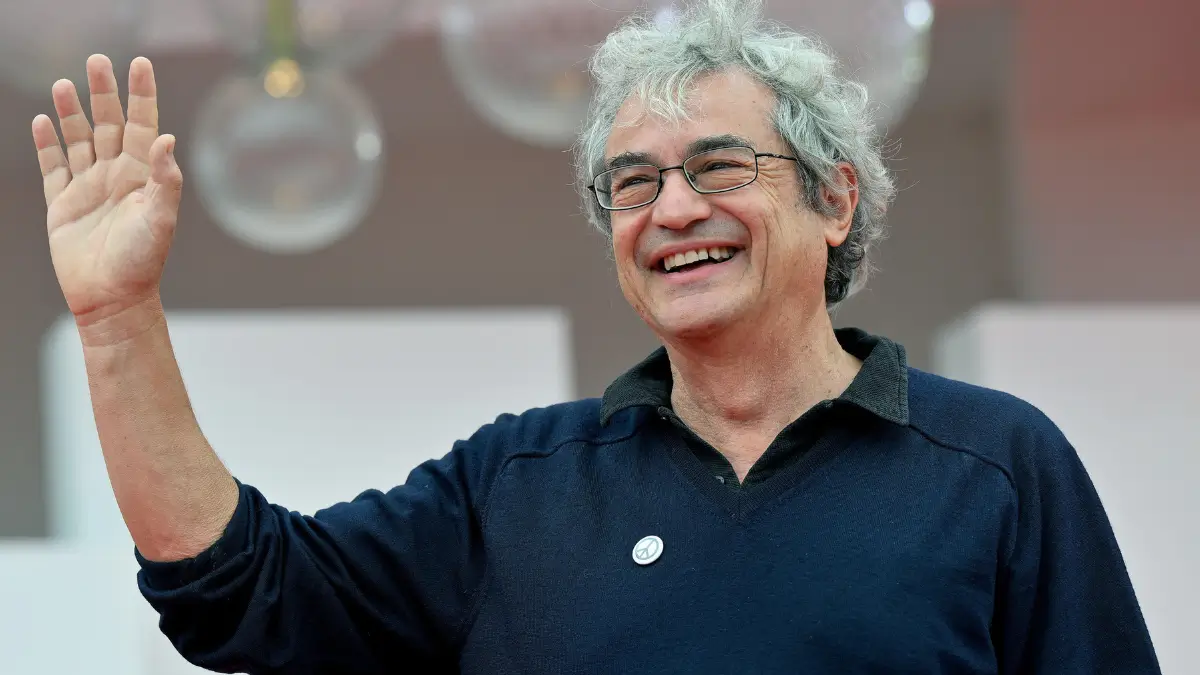 Carlo Rovelli, fisico e scrittore italiano, sorride e saluta con la mano destra alzata. Indossa un maglione scuro con colletto a polo nero e occhiali da vista. Ha capelli grigi arruffati