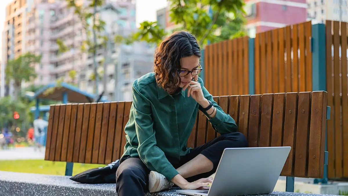 Una giovane donna con occhiali e camicia verde è seduta all'aperto su una panchina di legno e pietra, con un laptop sulle gambe. Sta guardando lo schermo e si tocca il mento con la mano, concentrata. Lo sfondo è un parco o una piazza urbana con alberi, vegetazione e un edificio in lontananza