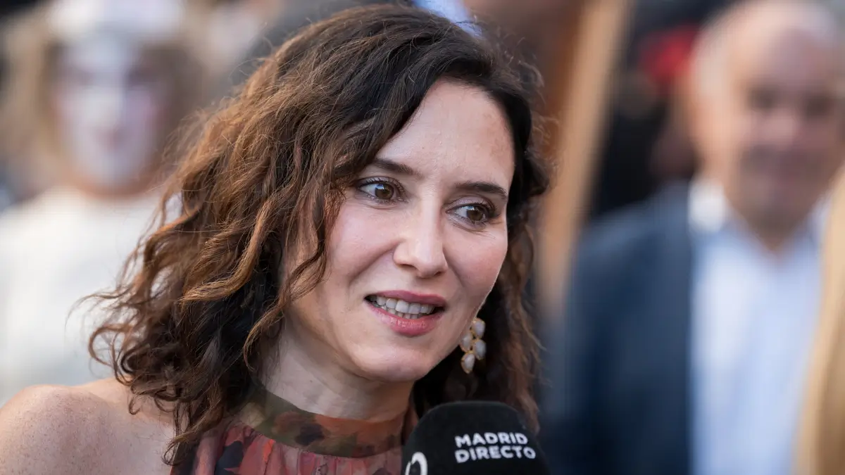 Isabel Díaz Ayuso, con capelli mossi castani e un abito floreale, parla a un microfono con la scritta “Madrid Directo” durante un’intervista all’aperto, con persone sfocate sullo sfondo