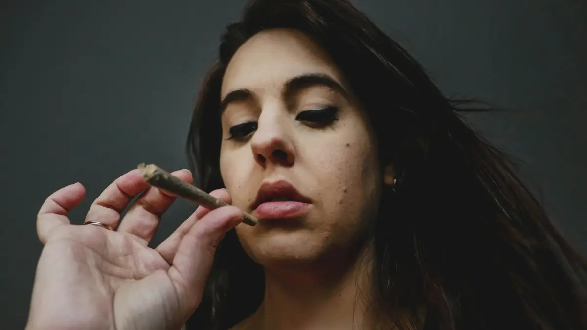 Donna con capelli scuri tiene una sigaretta arrotolata (che sembra essere una canna di marijuana) tra le dita vicino alla bocca, su uno sfondo grigio scuro