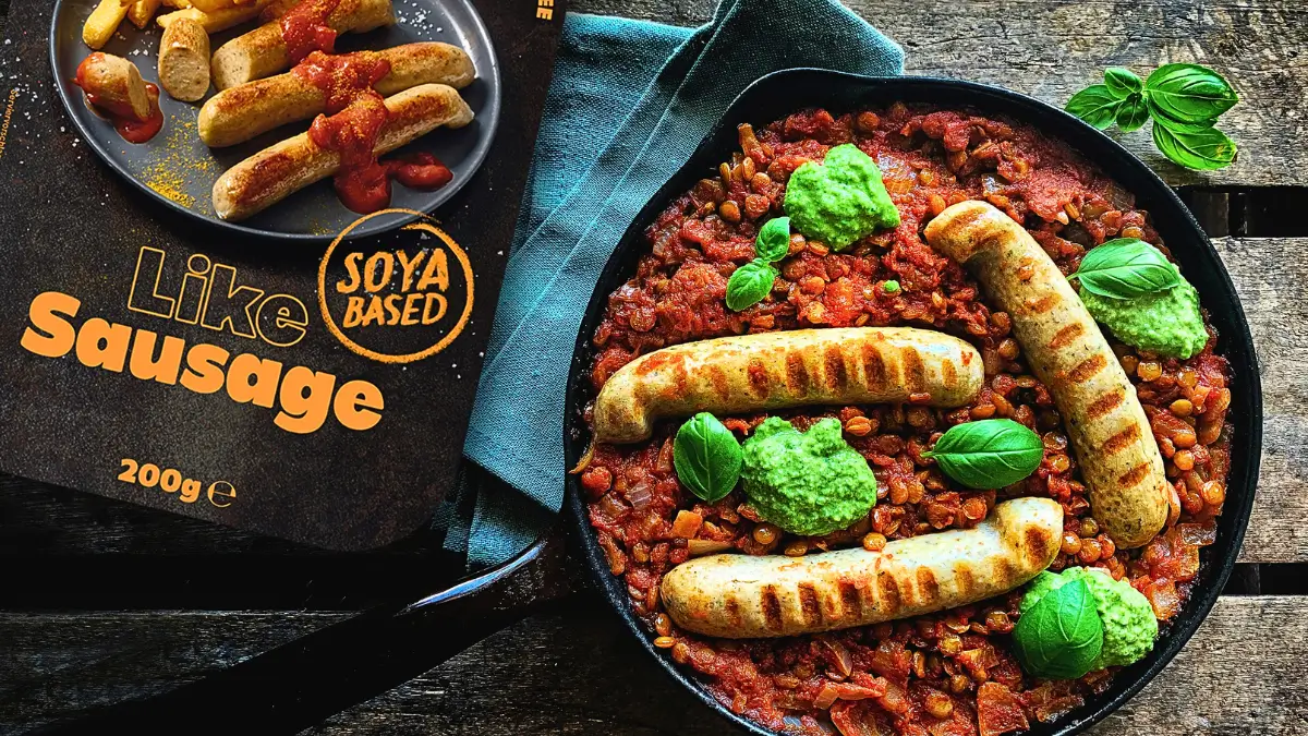 Vista dall'alto di una pietanza vegana che mostra tre salsicce grigliate a base di soia ('Like Sausage') servite su un letto di stufato di lenticchie e pomodoro. La pietanza è guarnita con ciuffi di salsa verde (come pesto o guacamole) e foglie fresche di basilico, ed è contenuta in una padella di ghisa nera. Accanto alla padella, c'è un piatto grigio con altre salsicce e patatine, oltre alla scatola del prodotto con il logo 'Soya Based'