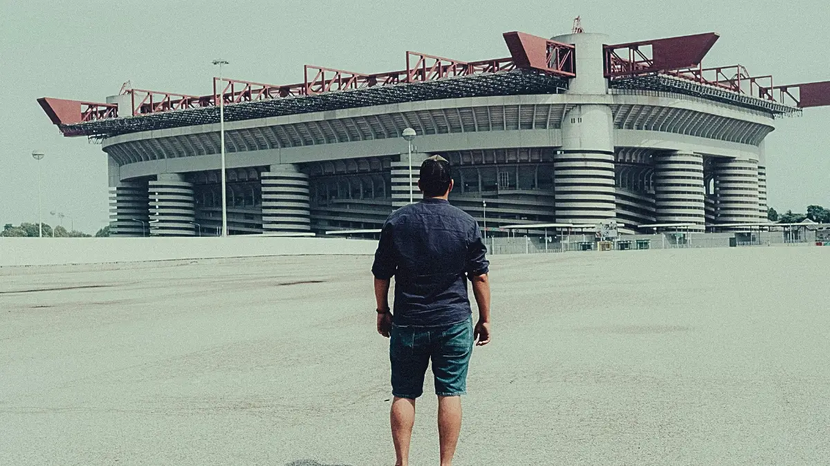 Un uomo visto di spalle, con maglietta blu scura e pantaloncini di jeans, si trova su una vasta spianata di cemento e guarda lo Stadio San Siro (Giuseppe Meazza) a Milano. L'iconica struttura dello stadio è visibile in primo piano con i suoi tre anelli distintivi e le torri esterne a spirale. L'immagine ha un tono retro o leggermente desaturato