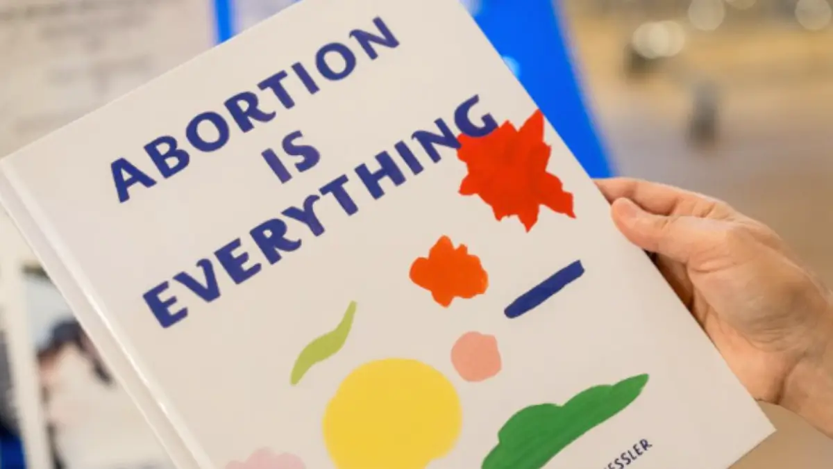 Copertina di un libro bianco per bambini tenuto da una mano. Il titolo è scritto in blu e recita: “ABORTION IS EVERYTHING”. La copertina è decorata con forme astratte e colorate che richiamano l'arte moderna o disegni per bambini (rosso, arancione, giallo, verde)