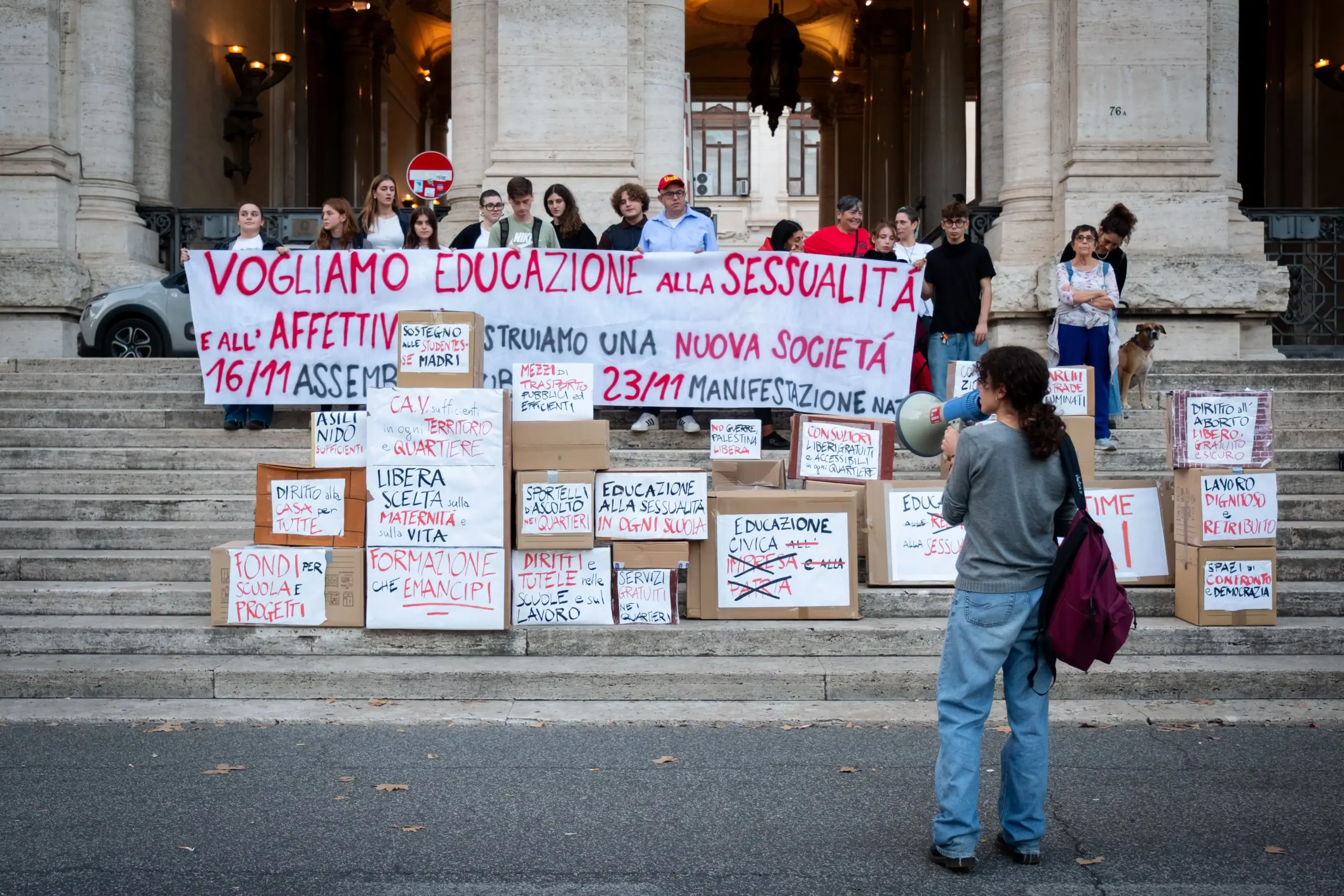 Manifestazione studentesca davanti a un grande edificio classico con scalinata. Un lungo striscione bianco recita in rosso: 'VOGLIAMO EDUCAZIONE ALLA SESSUALITÀ E AGLI AFFETTIVI. COSTRUIAMO UNA NUOVA SOCIETÀ'. Gli studenti sono in piedi dietro lo striscione. Sullo scalone sono impilati numerosi cartoni con scritte a mano che riportano varie richieste. Una manifestante in primo piano è rivolta di spalle e parla in un megafono