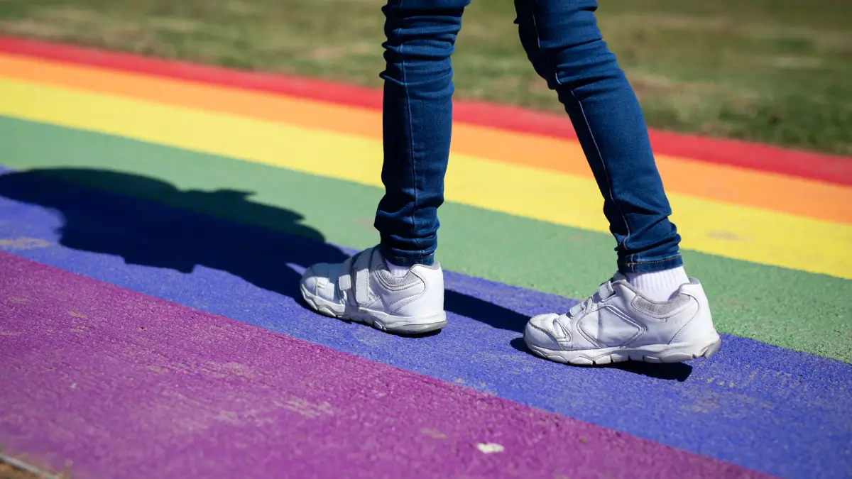 Persona con scarpe bianche e jeans che cammina sopra una bandiera arcobaleno LGBTQ+ dipinta a terra
