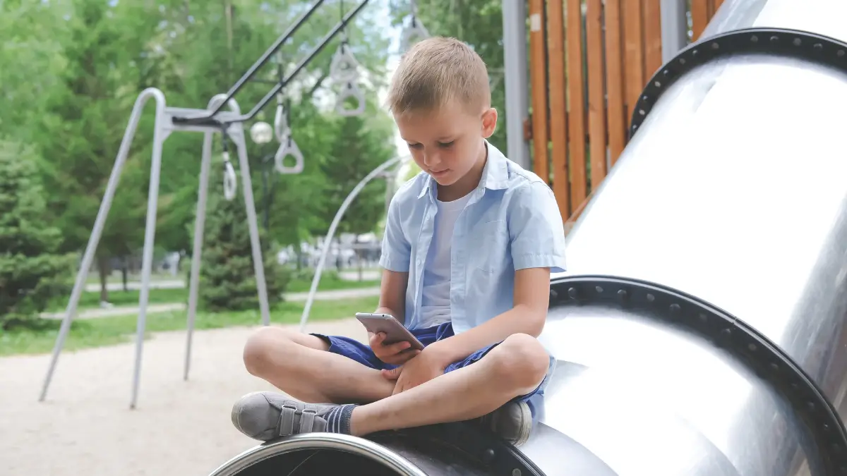 un bambino guarda uno smartphone seduto sullo scivolo di un parco giochi bambini social