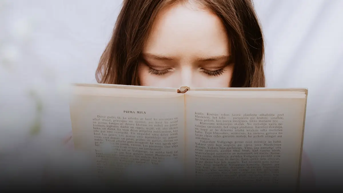 Primo piano del volto di una giovane ragazza con gli occhi socchiusi, parzialmente nascosto da un libro aperto che sta leggendo; lo sfondo è bianco e sfocato