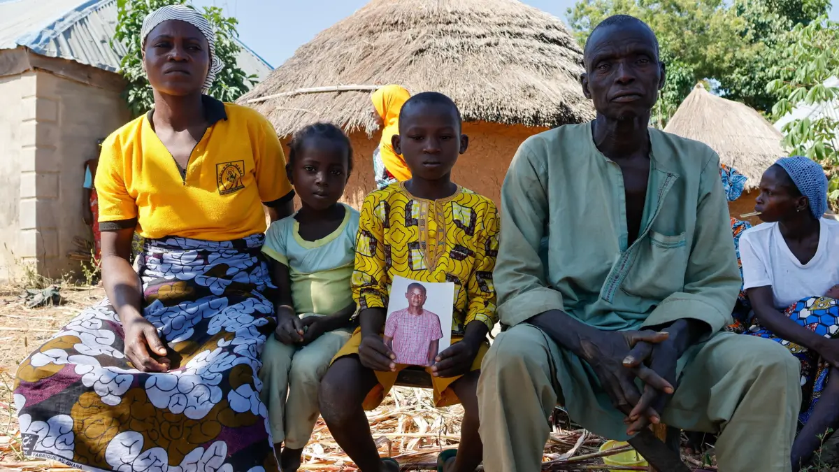 Una famiglia siede all'aperto davanti a capanne tradizionali in Nigeria; un bambino al centro tiene tra le mani la fotografia di un uomo giovane