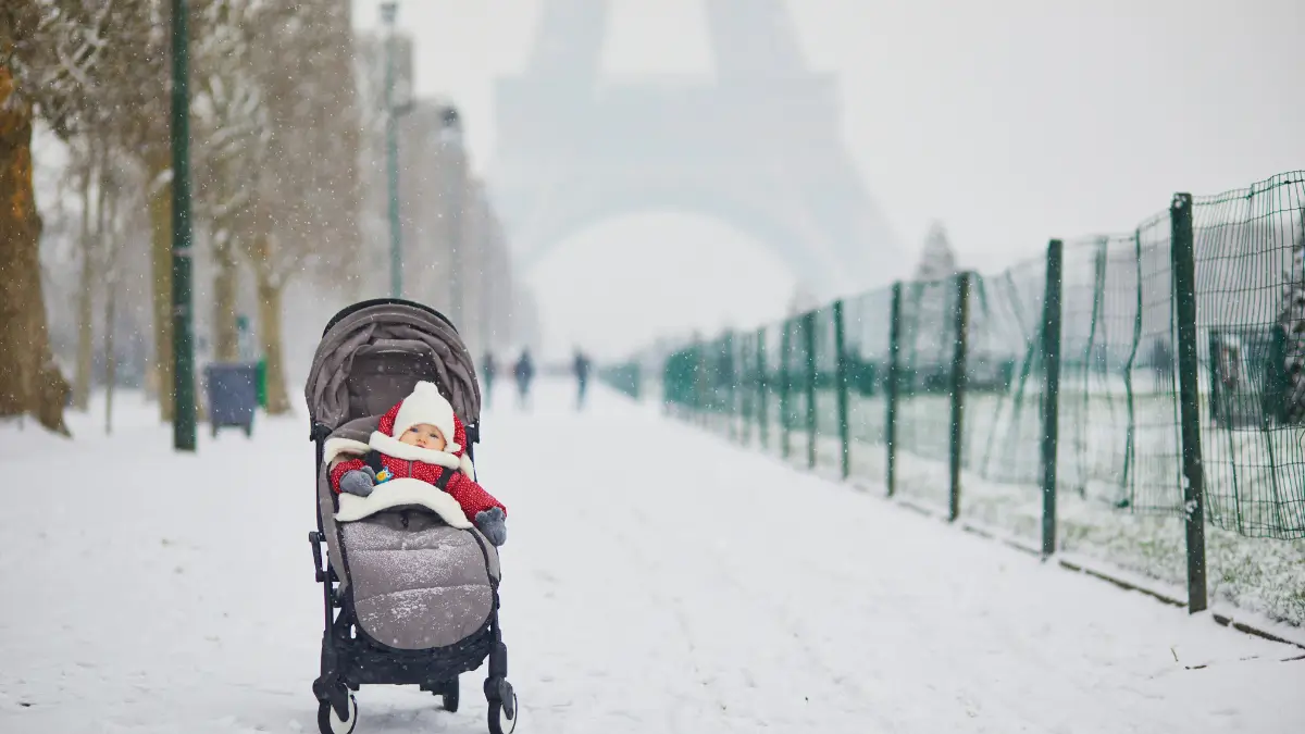 Un bambino piccolo avvolto in una tuta rossa e cappellino bianco siede in un passeggino grigio su un sentiero innevato a Parigi, con la sagoma sfocata della Torre Eiffel sullo sfondo