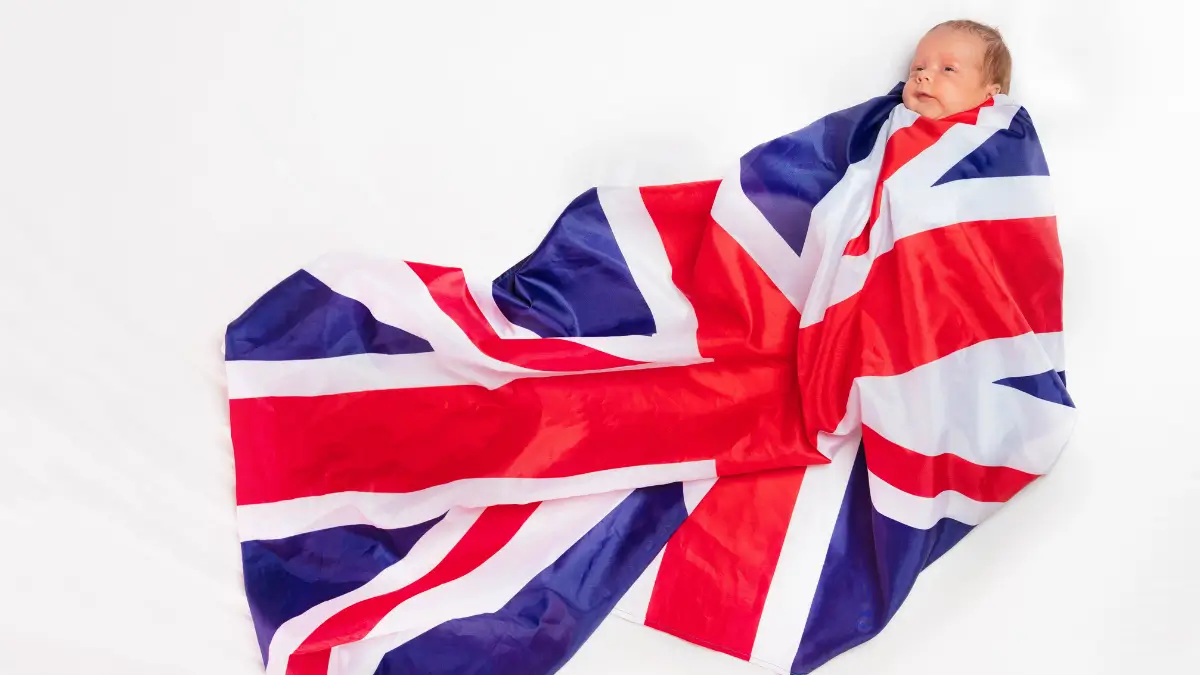 Un neonato disteso su una superficie bianca, avvolto interamente in una grande bandiera del Regno Unito (Union Jack) che si estende morbidamente intorno a lui