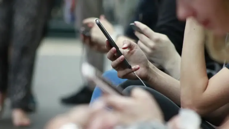 Primo piano delle mani di diverse persone sedute che tengono e utilizzano i propri smartphone; una mano al centro &egrave; a fuoco mentre tiene un telefono sottile collegato a degli auricolari bianchi.
