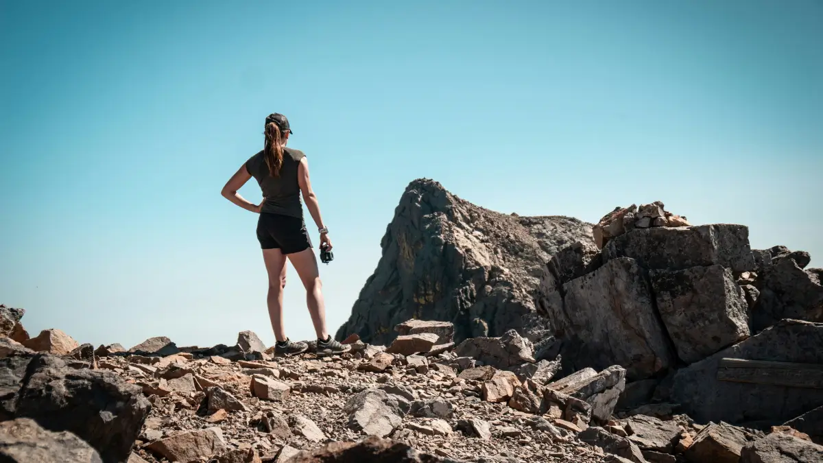 Una donna di spalle con abbigliamento sportivo e berretto osserva un'imponente vetta rocciosa sotto un cielo terso; si trova su un crinale sassoso e tiene una macchina fotografica in mano