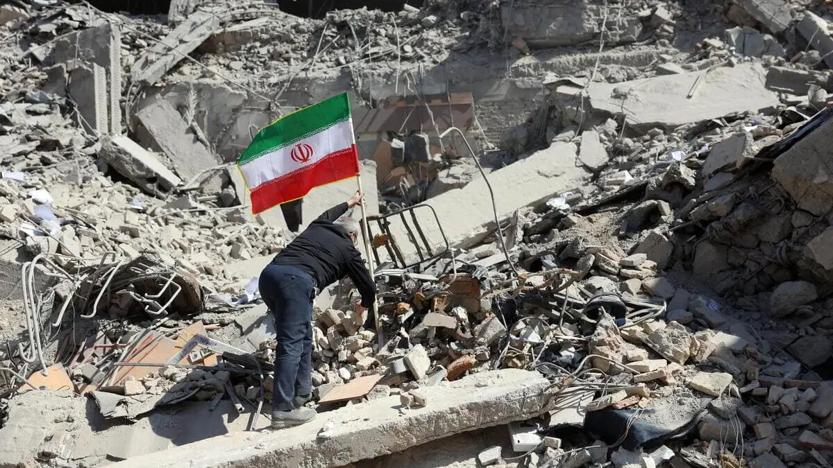 Un uomo pianta una bandiera dell'Iran su un cumulo di rovine dopo un bombardamento a Teheran