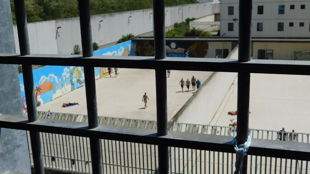 Veduta dall'alto di un cortile interno di un carcere attraverso le sbarre di una finestra; si vedono alcuni detenuti che camminano e muri perimetrali decorati con murales colorati