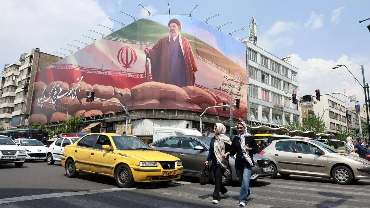 Due donne a Teheran, in Iran, attraversano la strada sotto un enorme cartello di propaganda per il regime islamico guidato da Mojtaba Khamenei